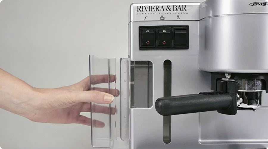 Une main retire le réservoir d'eau d'une machine à espresso Riviera & Bar argentée, les boutons de commande étant visibles sur le devant de la machine.