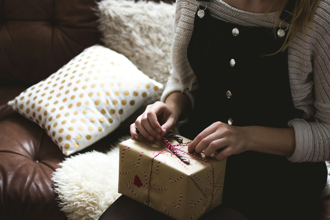 Une personne vêtue d'une robe noire à boutons blancs est assise sur un canapé et détache la ficelle d'un cadeau emballé dans du papier festif. Un oreiller blanc à pois dorés et un coussin moelleux se trouvent à côté d'eux.