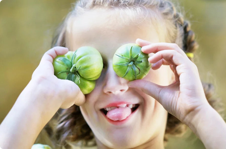 Un enfant souriant tient deux tomates vertes sur ses yeux et tire la langue de manière ludique, sur un fond vert flou.