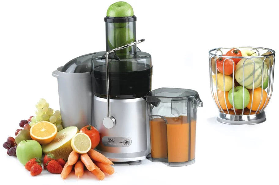 Un presse-agrumes argenté contenant du jus d'orange est posé sur un comptoir, entouré de fruits et légumes assortis, dont des carottes, des pommes, des oranges, des raisins et une corbeille de fruits entiers à l'arrière-plan.
