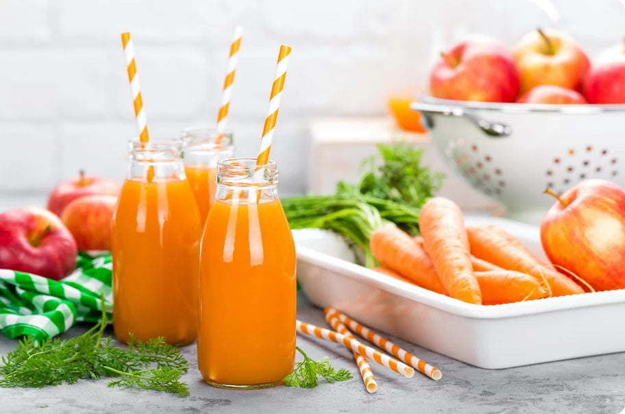 Trois bouteilles en verre remplies de jus d'orange et de pailles rayées sont placées à côté de carottes, de pommes et d'herbes fraîches sur un comptoir, avec des pommes supplémentaires dans une passoire blanche à l'arrière-plan.