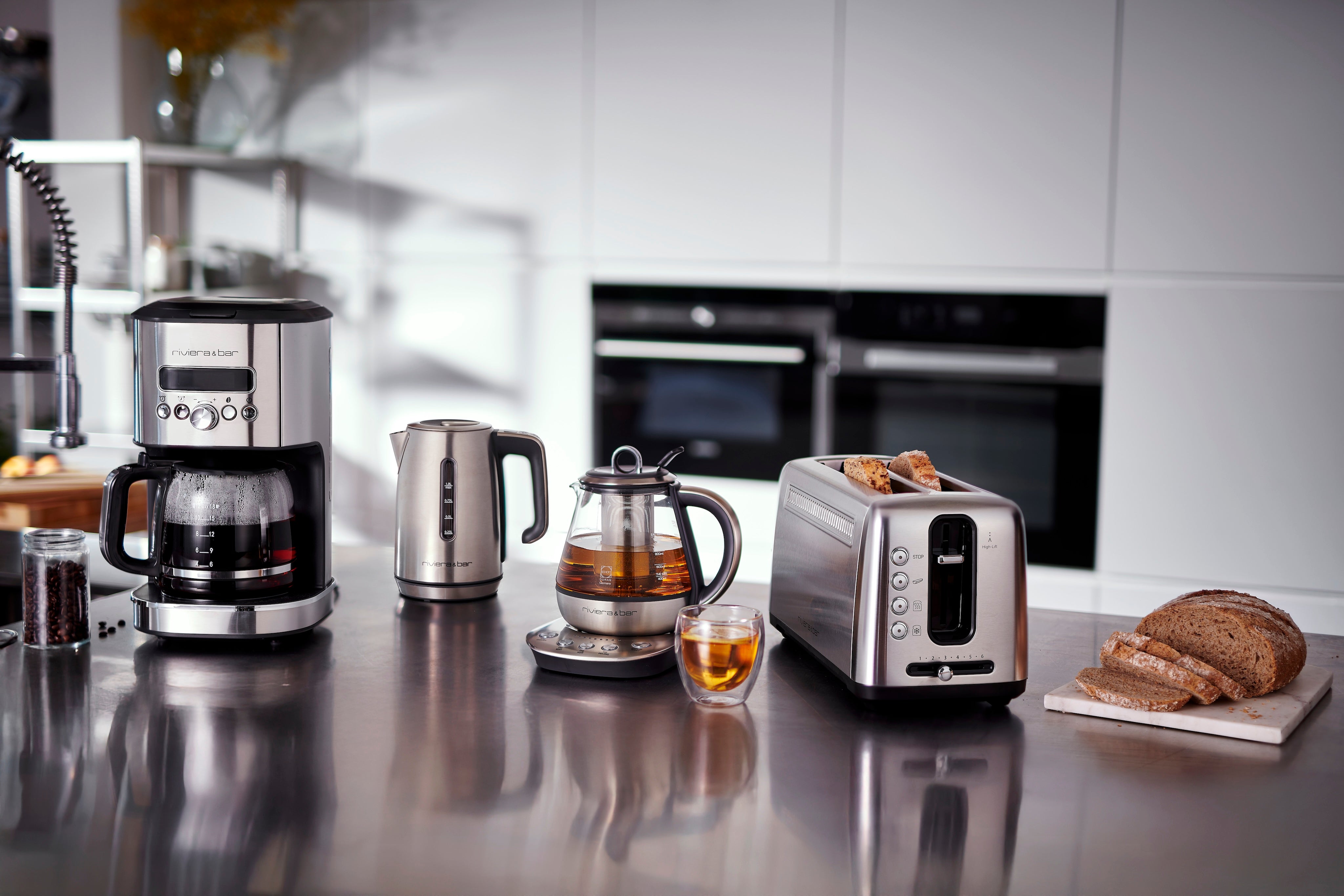 Petit accessoires électroménagers de la marque riviera&bar posés sur le plan de travail dans une cuisine moderne: une cafetière, une bouilloire à température réglable, une théière électrique et un grille-pain.