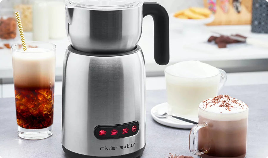 Un mousseur à lait électrique en acier inoxydable avec poignée noire est posé sur le comptoir d'une cuisine, entouré d'un verre de café glacé, d'une tasse de cappuccino au lait mousseux et d'une tasse de chocolat chaud garnie de copeaux de chocolat.