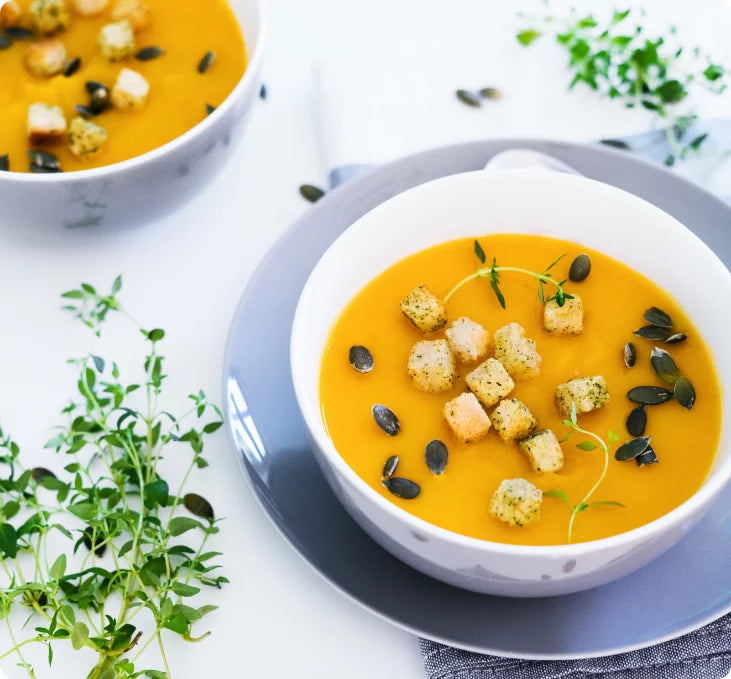 Un bol de soupe crémeuse orange garni de croûtons, de graines de citrouille et d'herbes fraîches est posé sur une assiette grise. Un deuxième bol et des brins d'herbes fraîches sont disposés à proximité sur une surface blanche.