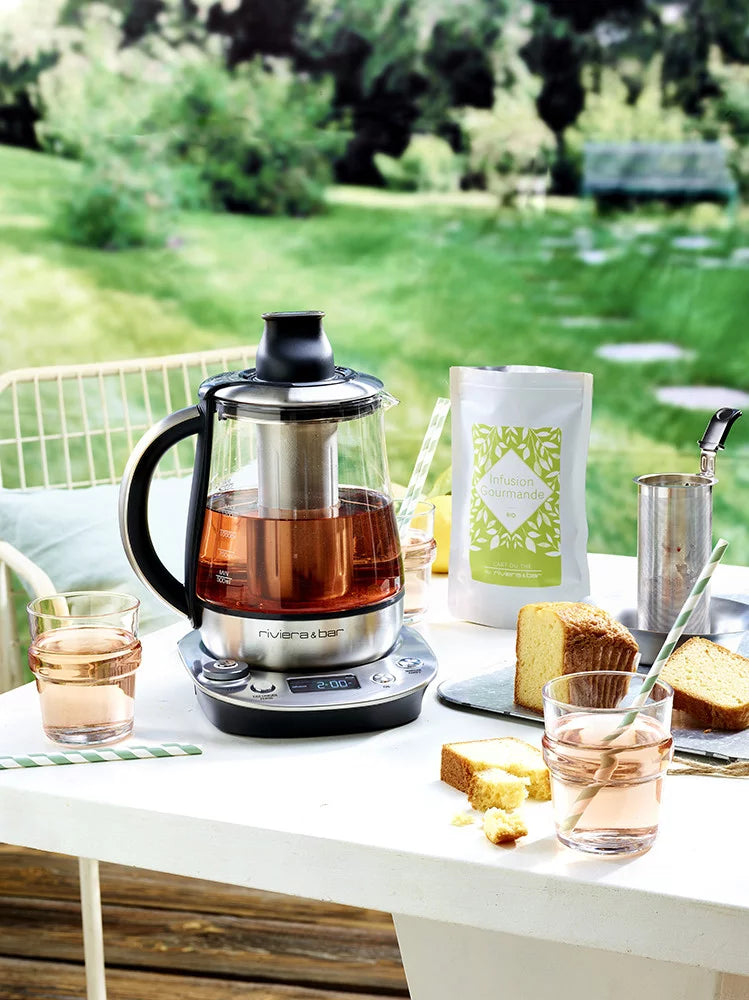 Une bouilloire électrique en verre avec du thé en train d'infuser est posée sur une table extérieure, entourée de tranches de gâteau, de verres de thé, d'un paquet de thé et d'un jardin verdoyant en arrière-plan.