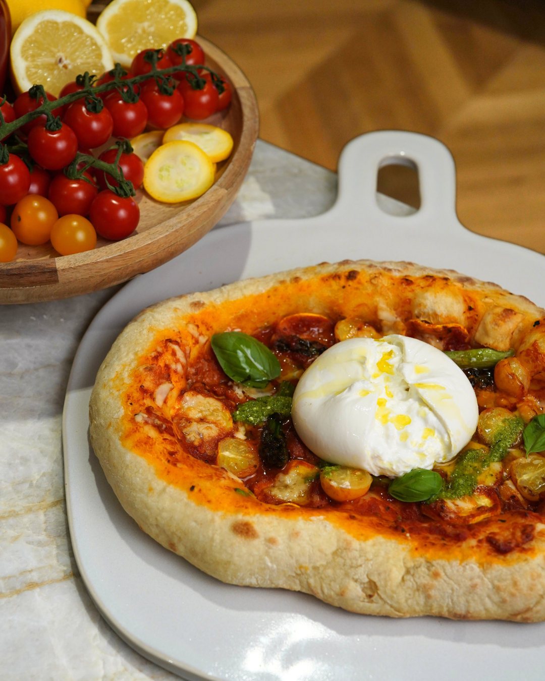 Une main soulève une part de pizza garnie de prosciutto, de mozzarella et de légumes dans une assiette. À proximité, des légumes grillés et deux salades sont disposés sur une table pour un repas.