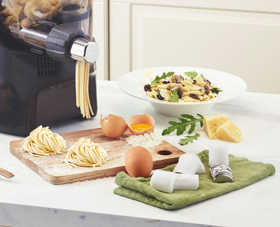 Une machine à pâtes extrude des pâtes fraîches sur une planche, à côté de laquelle se trouvent des œufs, de la farine et du fromage ; à l'arrière-plan, on aperçoit une assiette de pâtes cuites accompagnées de champignons et de roquette.