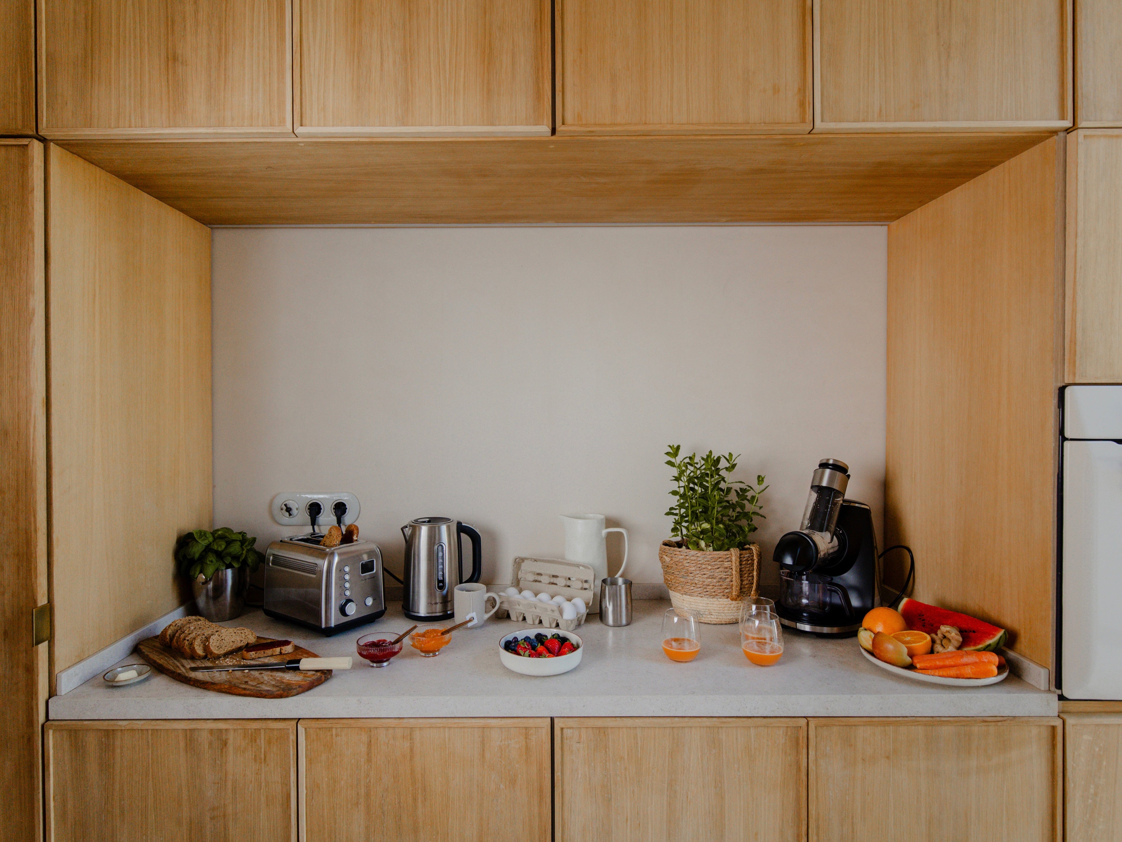 Un comptoir de cuisine moderne avec un grille-pain, une bouilloire, un mixeur, du pain frais, des fruits, du jus, des baies, une cafetière, une plante en pot et des verres, le tout posé sur des armoires en bois clair.