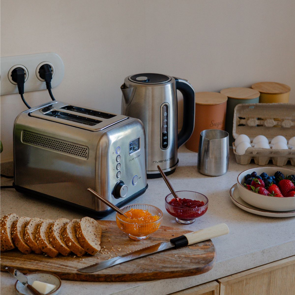 Un plan de travail en bois dans la cuisine, sur lequel sont disposés avec soin, contre un mur clair, du pain, un grille-pain, une bouilloire, des œufs, des baies, du jus de fruits, une machine à café, une plante en pot et des bols de fruits.
