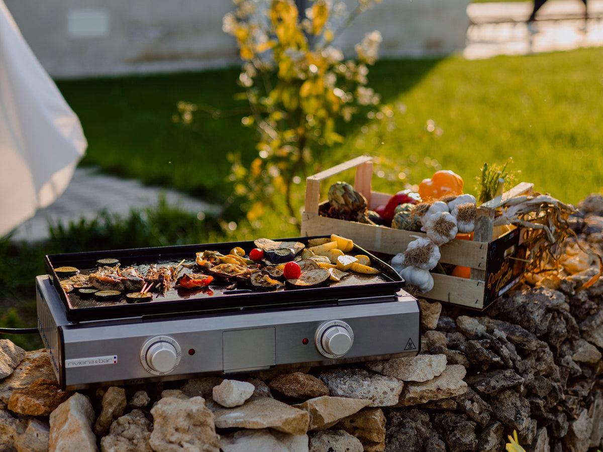 Au Riviera & Bar, un grill cuit des huîtres, du poisson et des légumes sur un mur de pierre, à côté d'une caisse de produits frais comme des artichauts et des poivrons.
