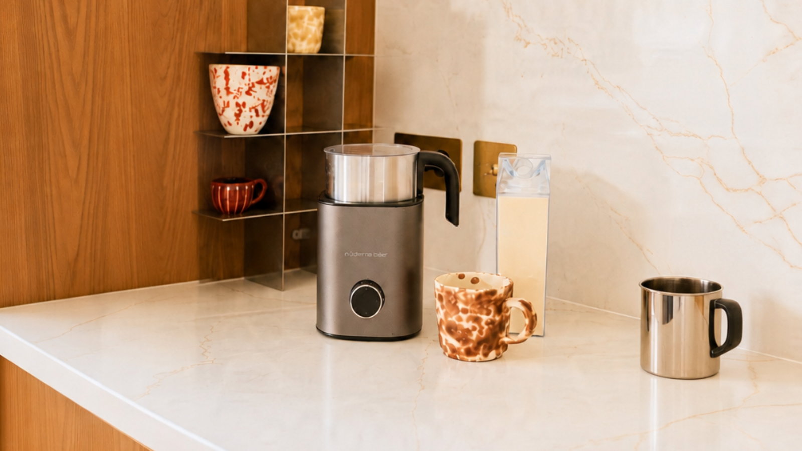 Un mousseur à lait, un mug en céramique à motifs, un carton de lait et un mug en inox posés sur un plan de travail en marbre.