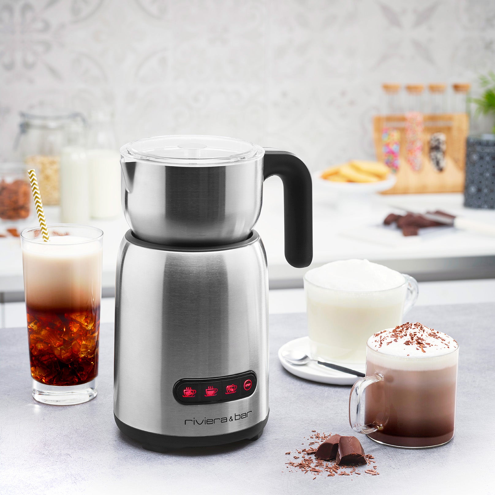 Un mousseur à lait en acier inoxydable avec une poignée noire est posé sur le comptoir d'une cuisine, entouré de boissons à base de café glacé, de cappuccino et de chocolat chaud. On aperçoit du lait, des morceaux de chocolat et un décor à l'arrière-plan.