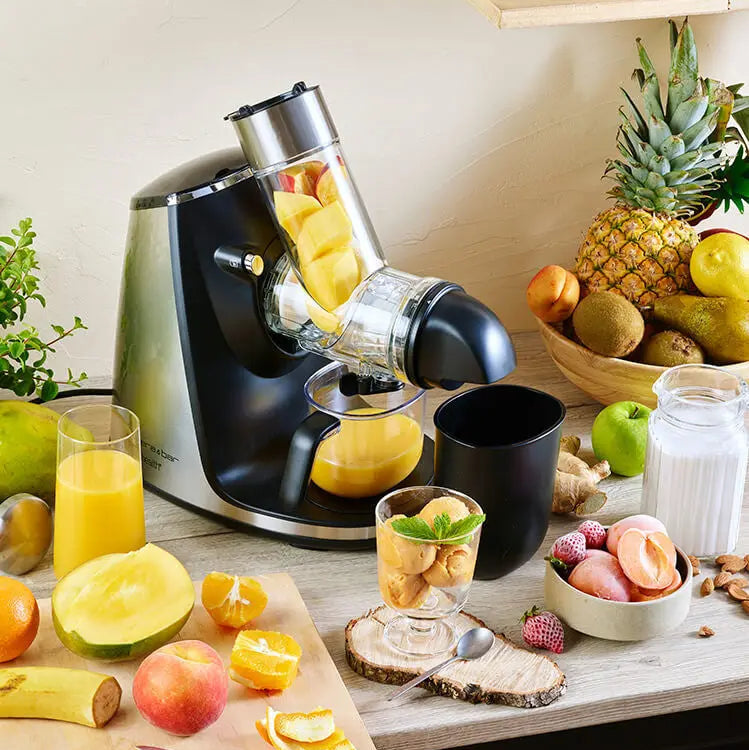 Gros plan d'un extracteur de jus en inox et noir, entouré de fruits frais et de récipients, sur une table en bois clair.
