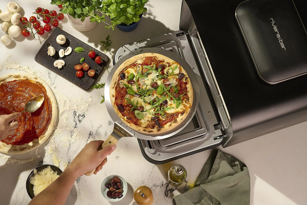 Quelqu'un glisse une pizza maison avec roquette et champignons dans un riviera&bar Four à pizza électrique 400 degrés, tandis que d'autres ingrédients et un autre plat italien sont préparés à proximité.