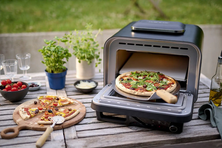 The four à pizza électrique 400 degrés de riviera&bar stoi sur une table en bois, cuisant une pizza maison. Autour : herbes, tomates cerise, verres, huile d'olive et une pizza tranchée avec coupe-pizza, dans un décor authentique.