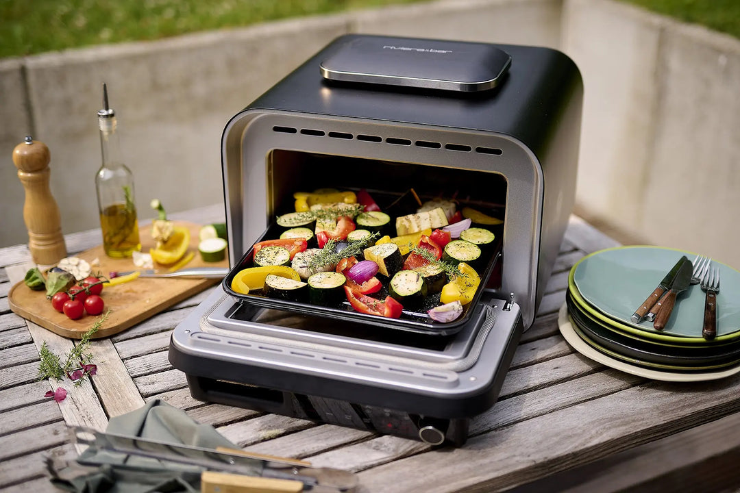 Le Four à pizza électrique 400 degrés de riviera&bar est posé sur une table de patio en bois avec des légumes tranchés, des assiettes, des ustensiles, de l'huile d'olive, un moulin à poivre, des tomates fraîches, du citron et des herbes - parfait pour un repas en plein air d'inspiration italienne.