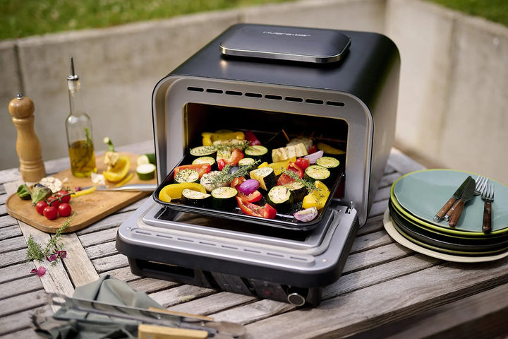 Le Four à pizza électrique 400 degrés de riviera&bar est posé sur une table de patio en bois avec des légumes tranchés, des assiettes, des ustensiles, de l'huile d'olive, un moulin à poivre, des tomates fraîches, du citron et des herbes - parfait pour un repas en plein air d'inspiration italienne.