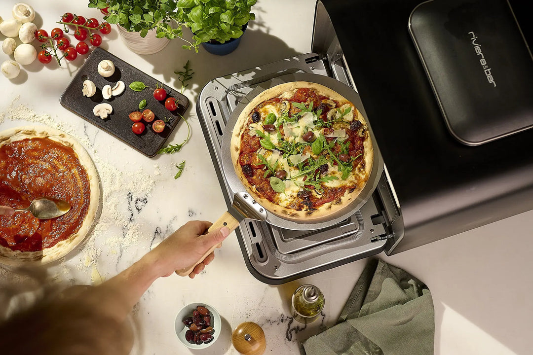 Une personne cuit une pizza maison fraîche garnie de légumes verts et de fromage dans le riviera&bar Four à pizza électrique 400 degrés, avec des tomates, des champignons et de la sauce sur le comptoir en marbre pour une véritable cuisine italienne.