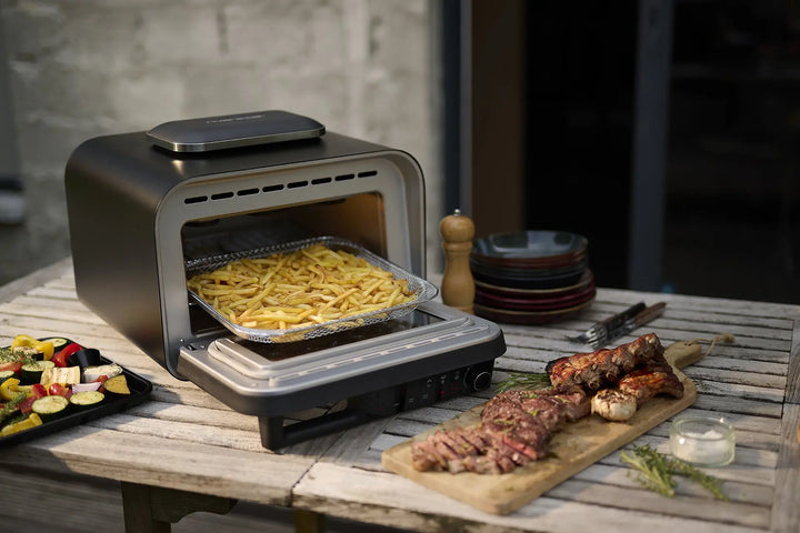 Un Four a pizza countertop oven by riviera&bar est posé sur une table d'extérieur, en train de cuire des frites. À proximité se trouvent une planche en bois avec de la viande grillée et des herbes, un bol de sauce, un moulin à poivre et une assiette de légumes grillés.