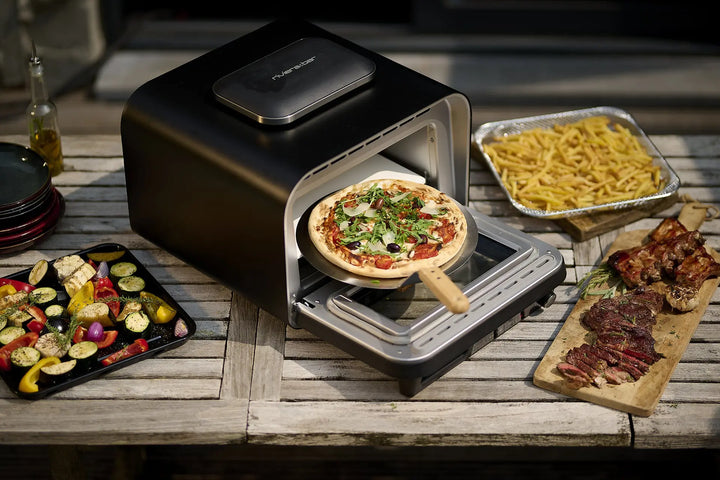 Le riviera&bar Un four à pizza cuit des pizzas sur une plaque en bois, avec des légumes rôtis, des pâtes, des viandes grillées et une bouteille d'huile exposée sur une table extérieure en bois à proximité.