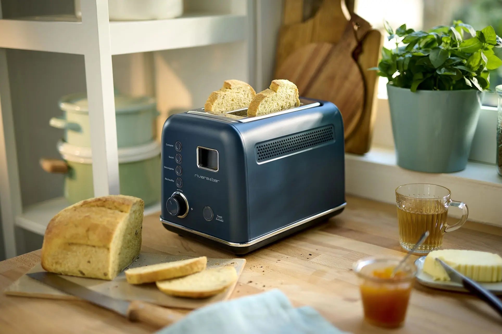 Une Grille-pain bleue riviera&bar avec éjection automatique est posée sur un comptoir de cuisine en bois à côté du pain, du thé, de la confiture, du beurre et d'une plante en pot. Profitez des neuf niveaux de stockage pour des toasts parfaits.