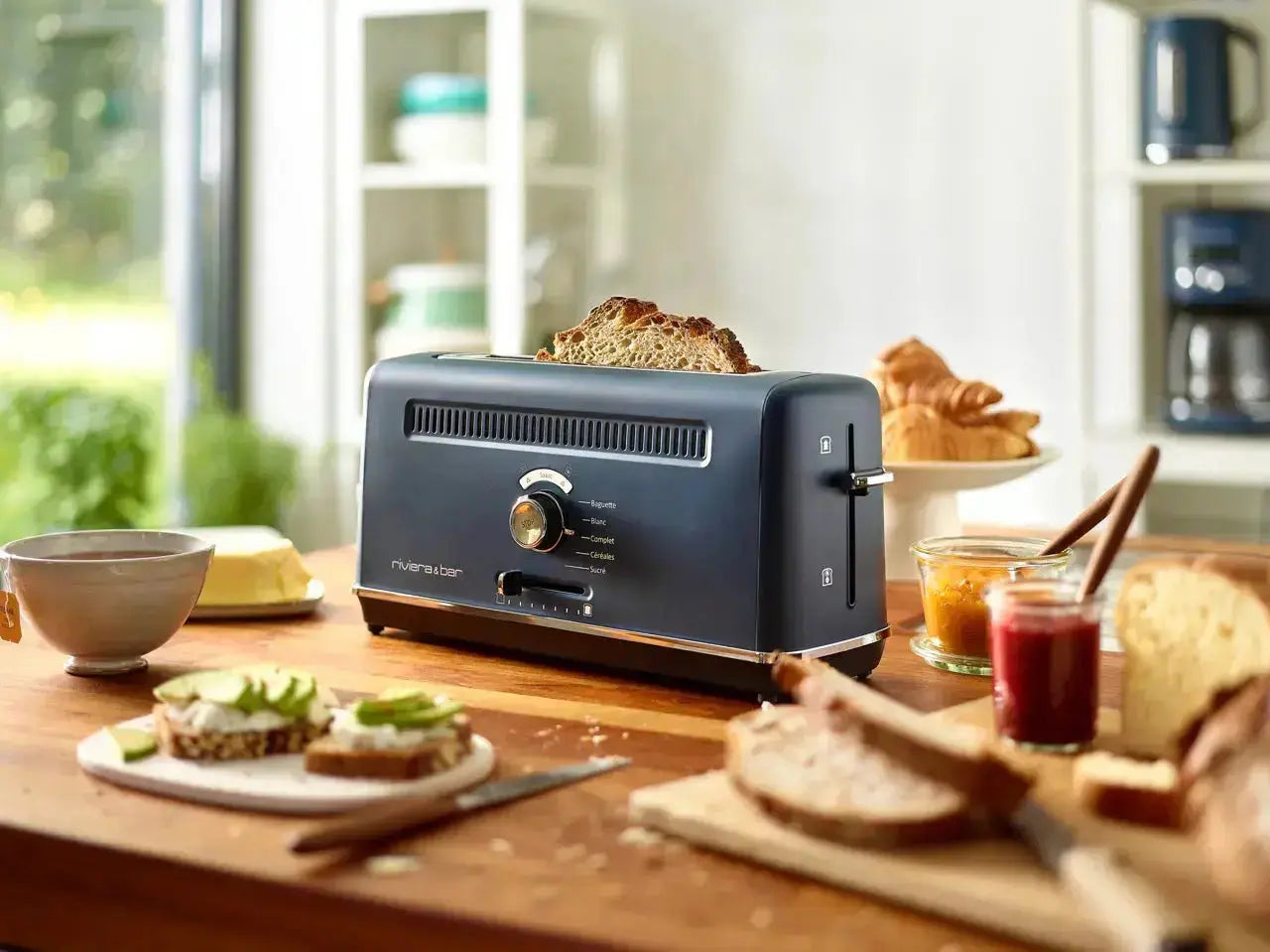 Un riviera&bar Ensemble petit-déjeuner vintage, avec un grille-pain bleu extra-long avec du pain à l'intérieur, est posé sur un comptoir en bois au milieu de tranches de pain, de beurre, de confiture et de toasts à l'avocat dans une cuisine lumineuse et éclairée par le soleil.