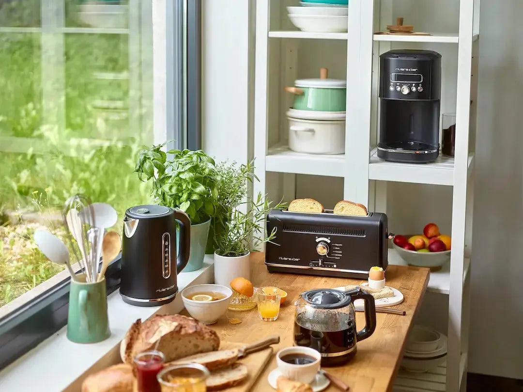 Une scène de cuisine lumineuse avec une table en bois dressée pour le petit-déjeuner, comprenant du pain, des fruits, du jus d'orange et du café. Sur le comptoir se trouve le riviera&bar Ensemble petit-déjeuner noir rétro complétant le look rétro.