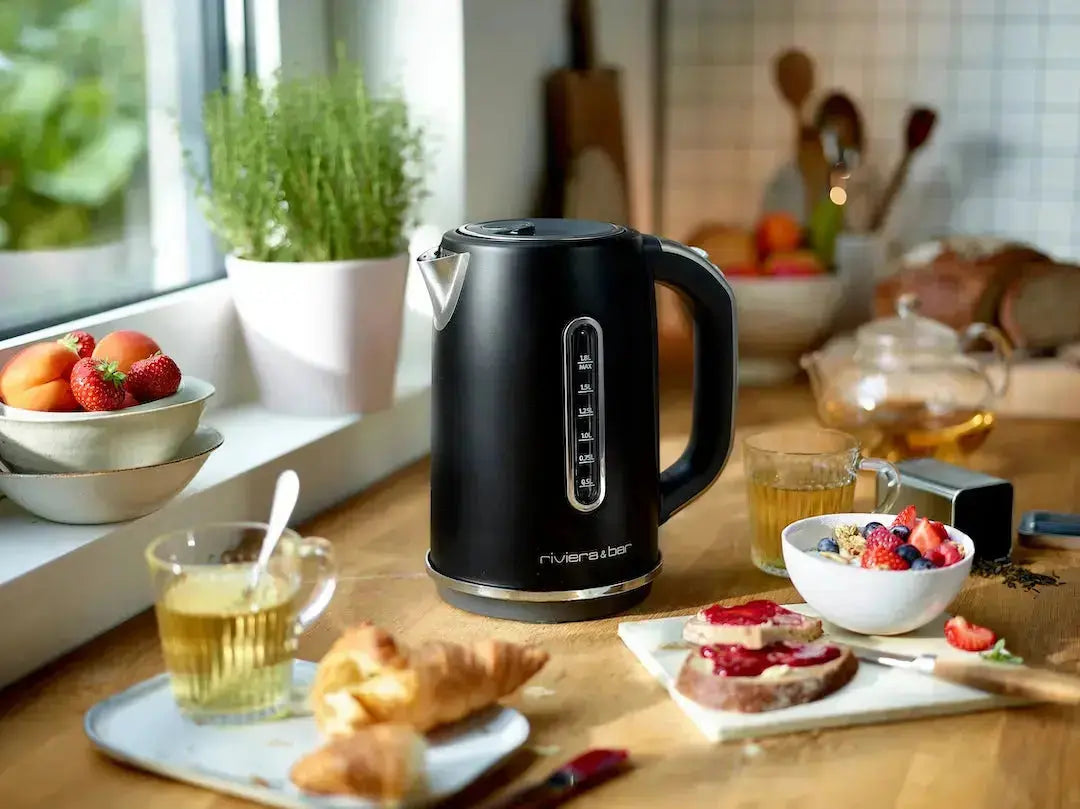 Une bouilloire Set grille-pain noir riviera&bar repose sur un comptoir en bois, présentée dans une charmante scène de petit-déjeuner avec thé, toast et confiture, croissant, coupe de fruits, tasse de thé, entourée de plantes et de pain.