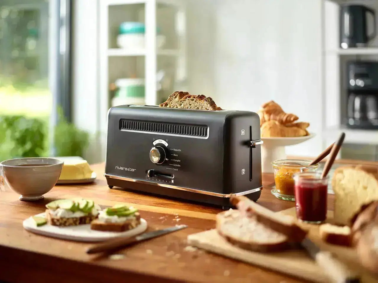 Une bouilloire Set grille-pain noir de riviera&bar est posée sur un comptoir de cuisine en bois, entourée de produits de petit-déjeuner, de pain, de bols et d'assiettes, tandis que la lumière du soleil pénètre par la fenêtre.