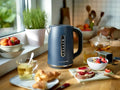 Une bouilloire bleue Set grille-pain vintage de riviera&bar est posée sur un comptoir de cuisine en bois près de tasses de thé, d'un petit-déjeuner composé d'un croissant, de toasts avec de la confiture, de fruits et d'herbes fraîches. La lumière du soleil entre par la fenêtre.