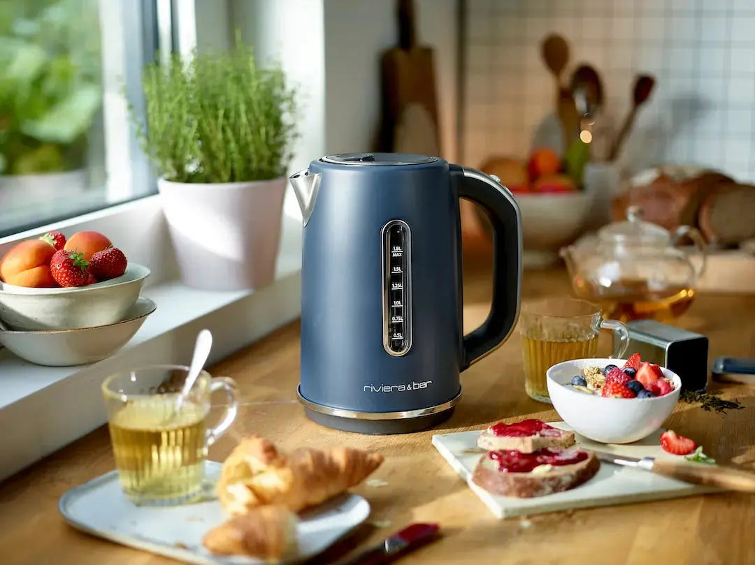 Une bouilloire bleue Set grille-pain vintage de riviera&bar est posée sur un comptoir de cuisine en bois près de tasses de thé, d'un petit-déjeuner composé d'un croissant, de toasts avec de la confiture, de fruits et d'herbes fraîches. La lumière du soleil entre par la fenêtre.