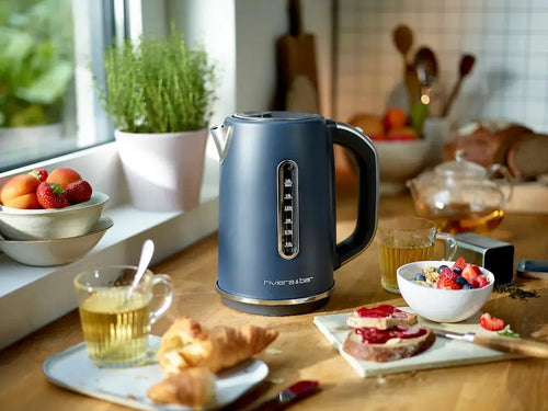 Une bouilloire bleue Set grille-pain vintage de riviera&bar est posée sur un comptoir de cuisine en bois près de tasses de thé, d'un petit-déjeuner composé d'un croissant, de toasts avec de la confiture, de fruits et d'herbes fraîches. La lumière du soleil entre par la fenêtre.