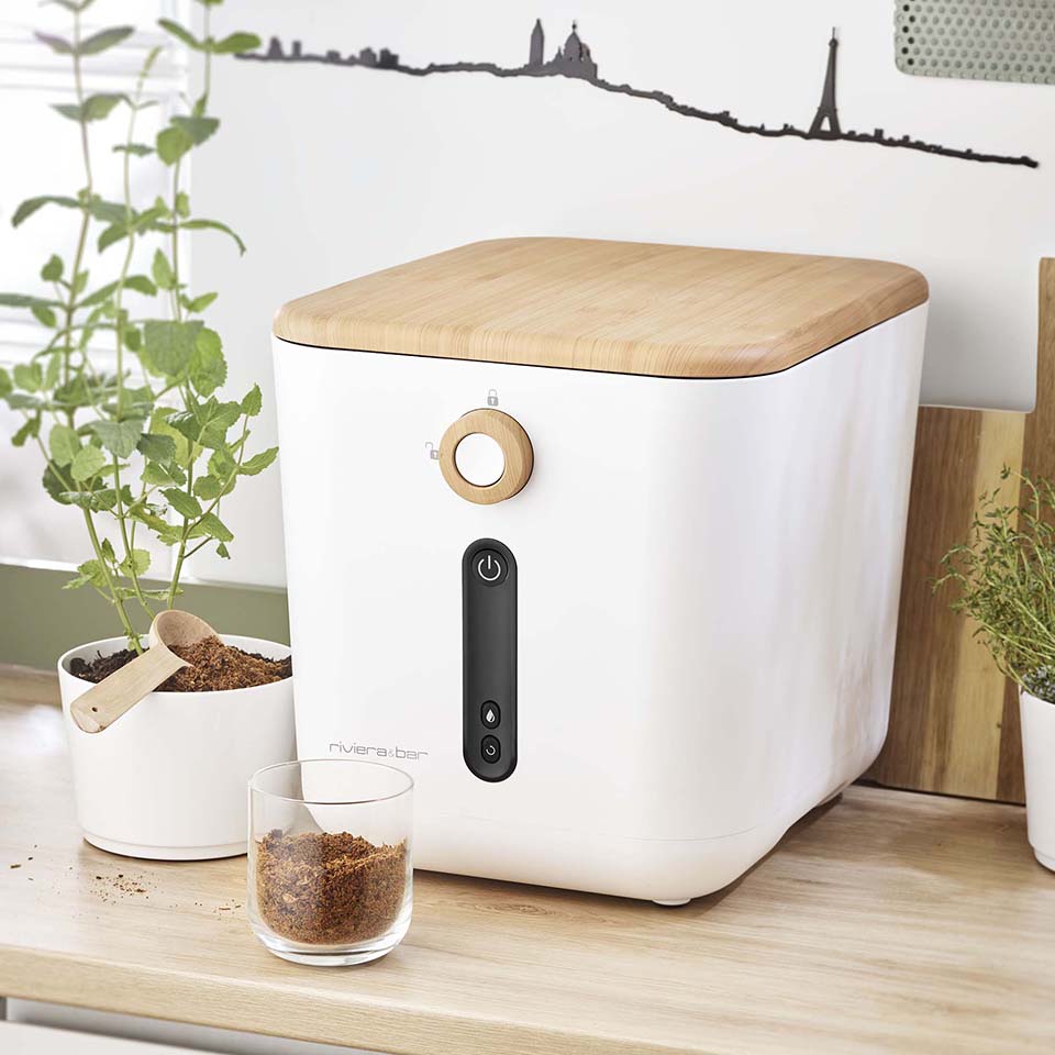Un appareil de cuisine blanc en forme de cube avec un couvercle en bois est posé sur un comptoir à côté d'herbes fraîches. Un verre contenant des granulés bruns et une cuillère en bois se trouvent au premier plan. Un tableau mural représentant l'horizon de Paris décore l'arrière-plan.