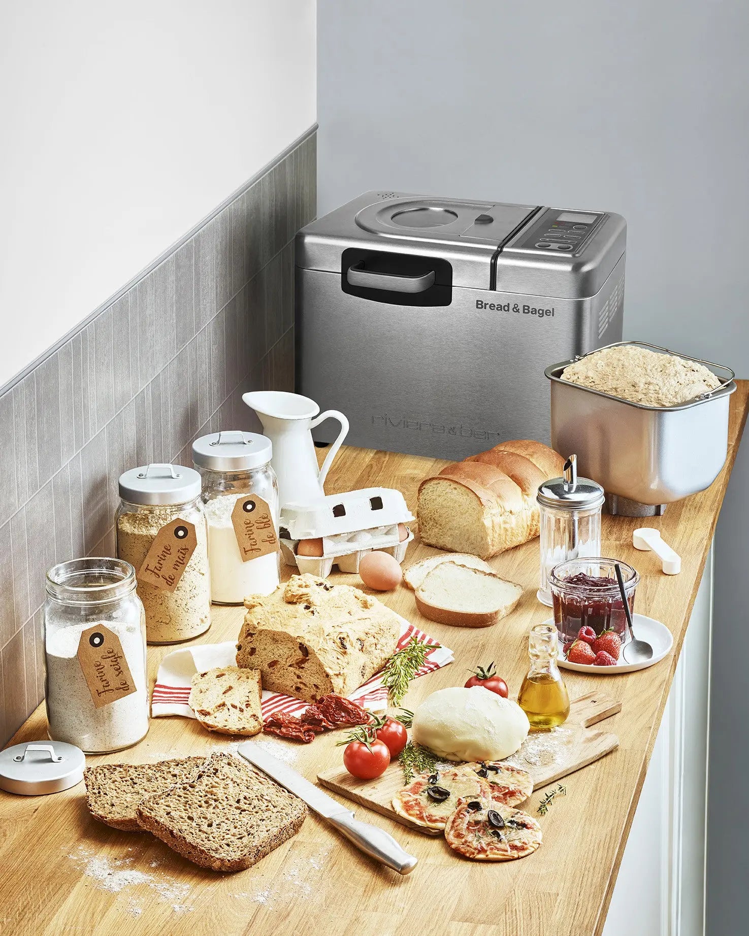 Voici quelques balises alt possibles en français pour cette image :

* Une machine à pain argentée sur un comptoir en bois, entourée de farine, de pain, de confiture, d'œufs et d'autres ingrédients de cuisine.
* Une image de produits mettant en scène une machine à pain, des ingrédients et des produits de boulangerie.
* Une nature morte de produits de boulangerie, avec une machine à pain, des pots de farine, du pain, de la confiture et d'autres ingrédients sur un comptoir en bois.
