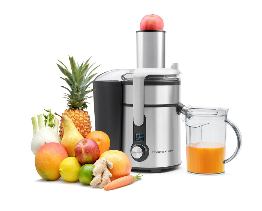 Un presse-agrumes en acier inoxydable avec une pomme à l'intérieur se trouve à côté d'un pichet en verre rempli de jus d'orange et d'une variété de fruits et légumes frais, dont un ananas, une pomme, des poires, des agrumes, du gingembre, une carotte et du fenouil.