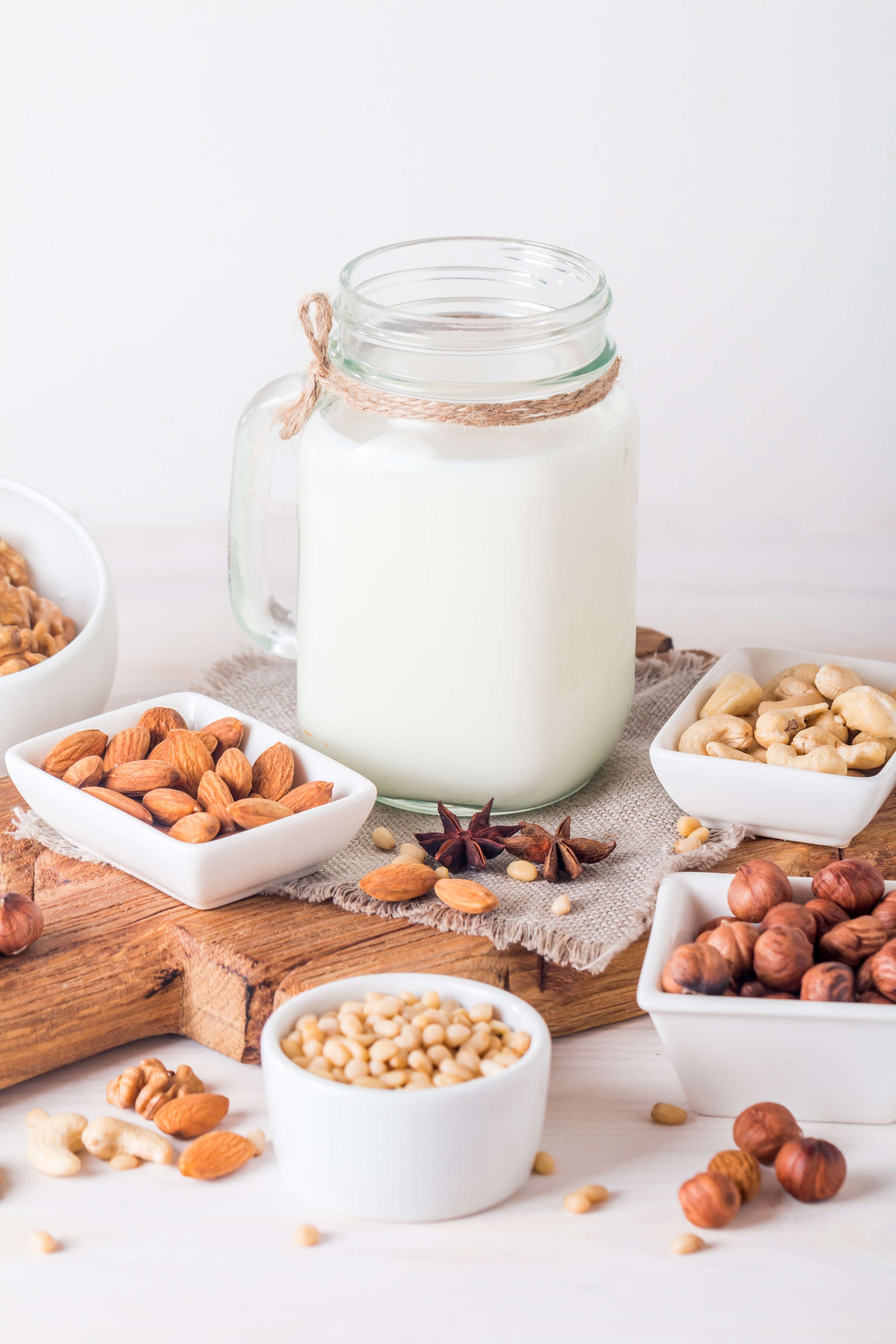 Un pot de lait en verre est posé sur une planche en bois, entouré de petits bols contenant divers fruits à coque, dont des amandes, des noisettes, des noix, des pignons et des noix de cajou. La scène a un aspect léger et rustique.