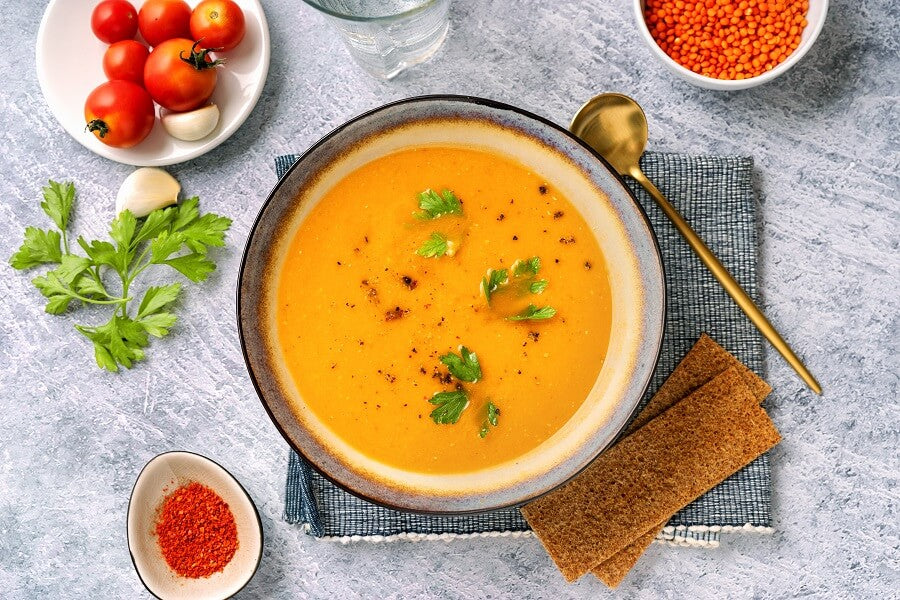 Un bol de soupe orange crémeuse garnie de persil est posé sur une serviette, avec une cuillère en or à proximité, entouré de pain croustillant, de tomates cerises, d'épices, de lentilles rouges et d'un verre d'eau sur une surface claire.
