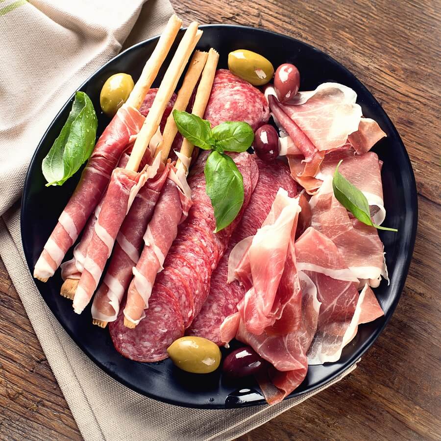 Une assiette noire avec un assortiment de charcuteries, des gressins enveloppés de prosciutto, des olives vertes et noires et des feuilles de basilic frais sur une table en bois avec une serviette beige.