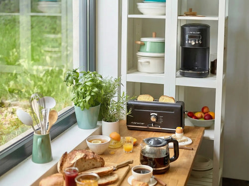 Une scène chaleureuse dans la cuisine, avec tout le nécessaire pour le petit-déjeuner — pain, café, jus de fruits et fruits disposés sur une table en bois près de la fenêtre —, met en valeur la cafetière et le grille-pain « Duo » de riviera&bar | Noir satiné, accompagnés de pots d'herbes aromatiques. En arrière-plan, des étagères garnies de vaisselle et de bols viennent compléter ce décor de petit-déjeuner accueillant.