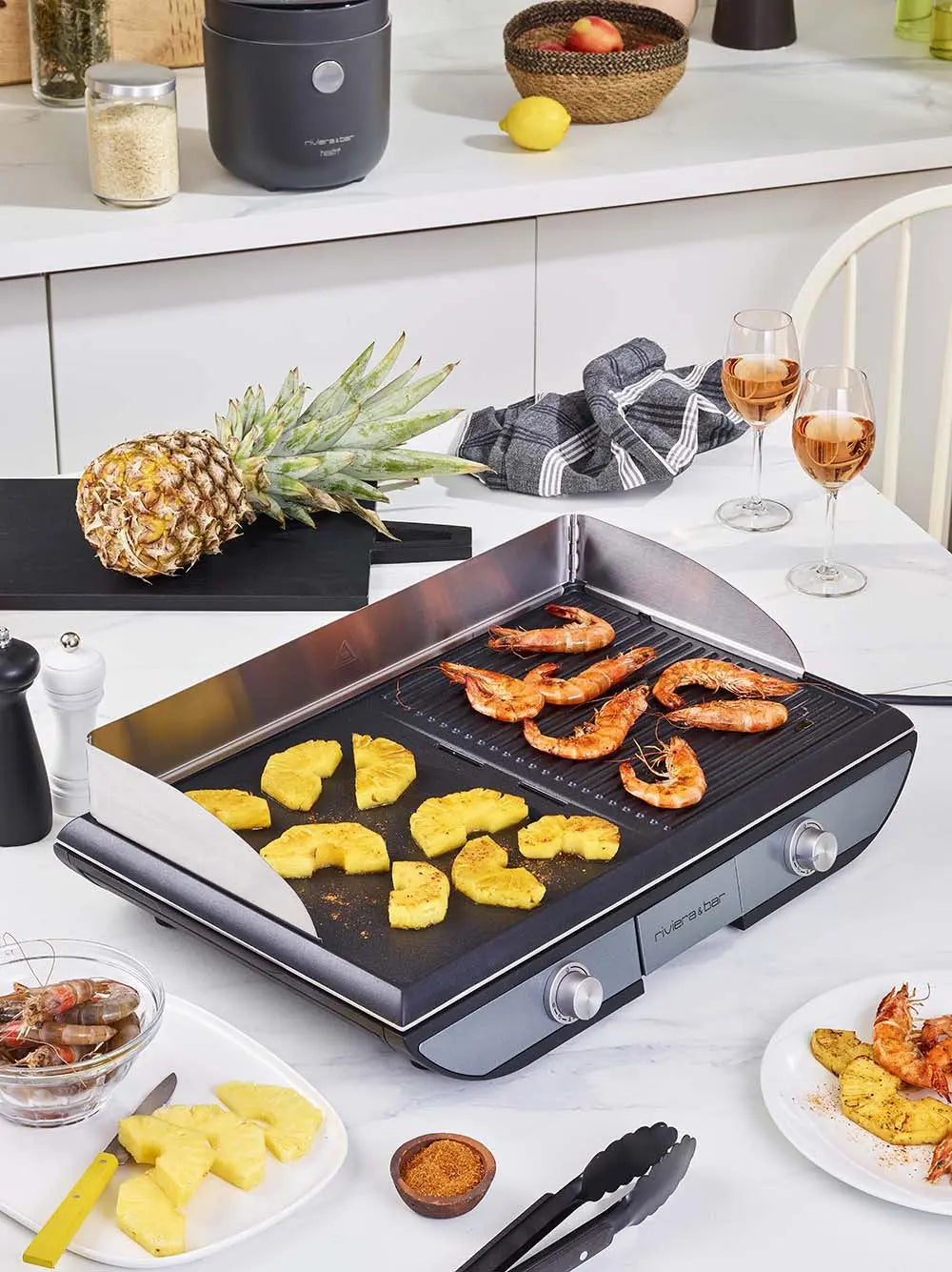 Un comptoir de cuisine présente la Plancha 2 zones de cuisson de riviera&bar avec une surface antiadhésive, grillant des ananas et des crevettes. À proximité se trouvent un ananas frais, un bol de crevettes, des verres à vin rosé et des ustensiles de cuisine tels qu'un cuiseur à riz et un moulin à poivre.