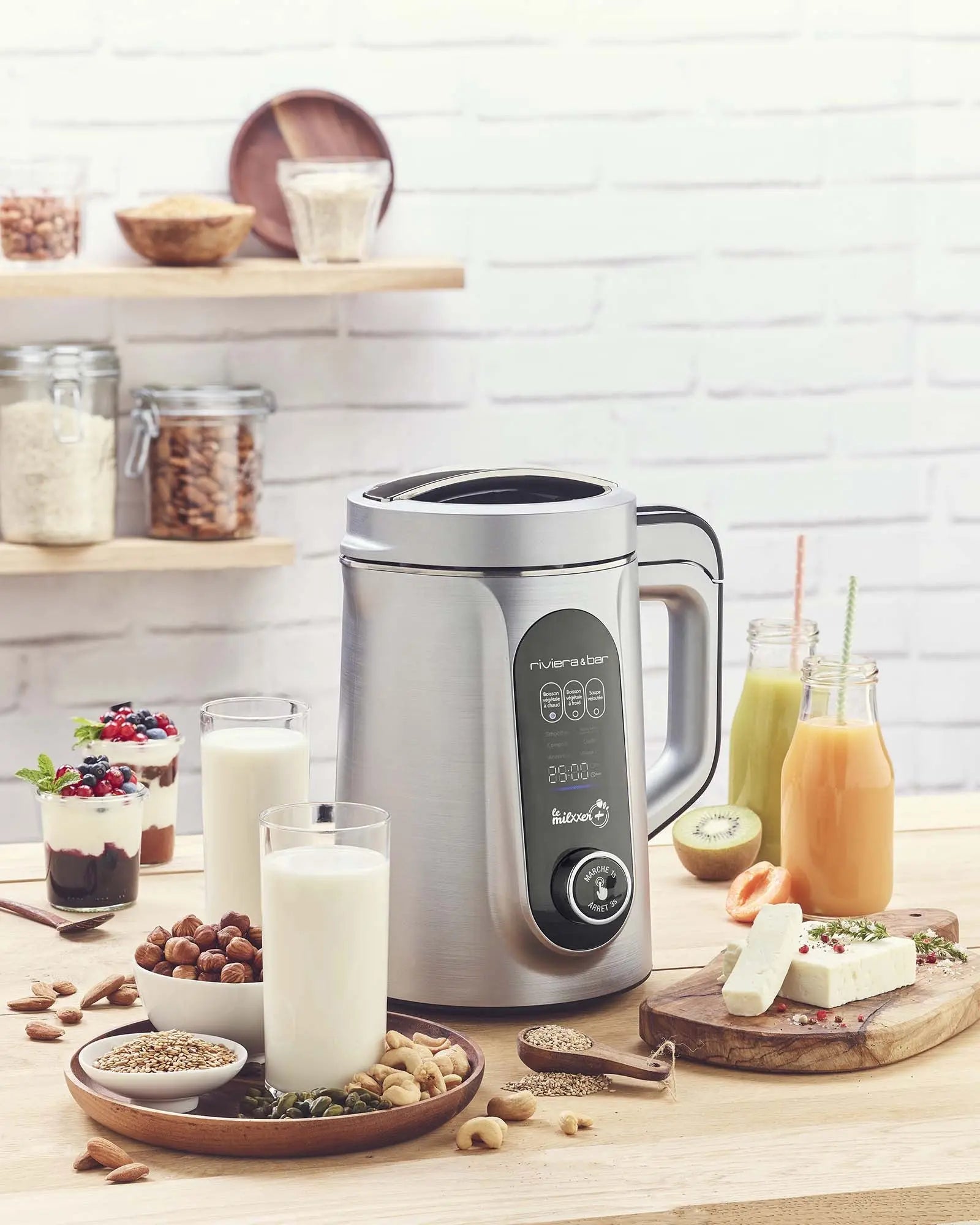 La machine à boissons en acier inoxydable riviera&bar Milxxer+ est posée sur un comptoir en bois avec des verres de lait, un assortiment de noix, de graines, de fruits et de barres dessert, tandis que d'autres ingrédients et bocaux sont alignés sur les étagères contre un mur de briques blanches.