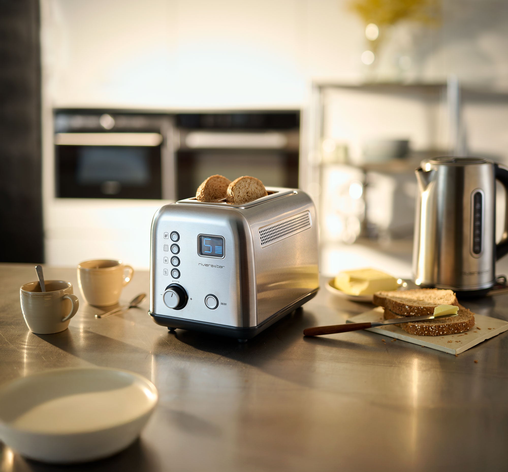 Un riviera&bar Grille-pain automatique en inox, avec écran LCD pour un brunissement parfait, est posé sur un comptoir de cuisine à côté d'une bouilloire, de tasses à café, de pain beurré, d'un beurrier et d'un couteau dans un environnement doucement éclairé.