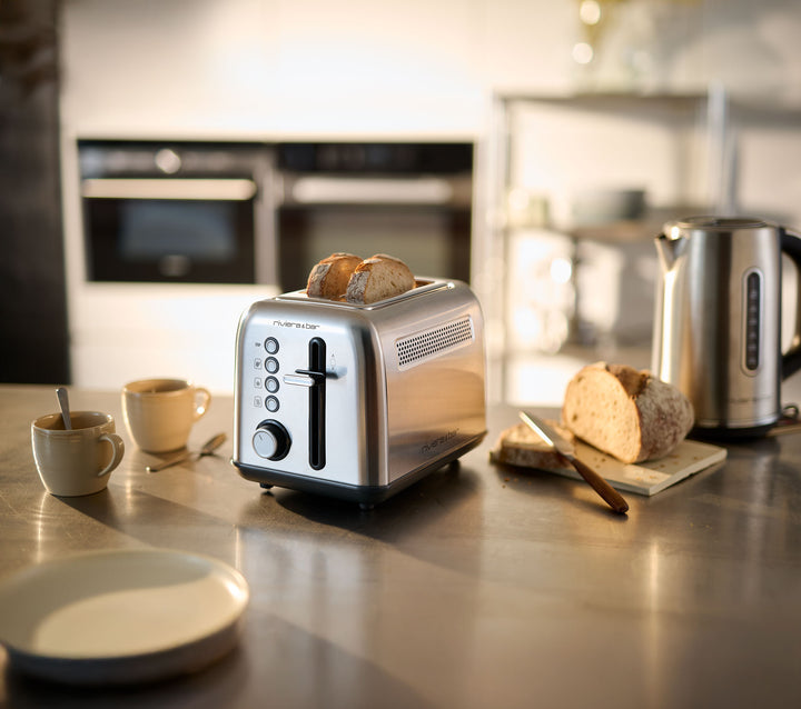 La grille-pain inox QGP220 de riviera&bar à 2 fentes courtes accueille deux tranches sur un plan de travail, près d'une bouilloire, d'un pain, d'un couteau et de deux tasses dans une cuisine chaleureusement éclairée.