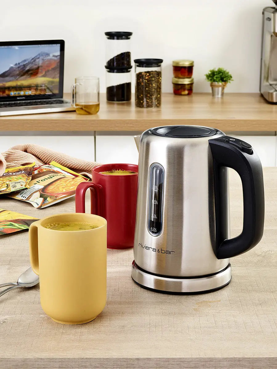 La Bouilloire Sofia - Inox (1 L) de riviera&bar est posée sur le comptoir de la cuisine à côté d'une tasse jaune et rouge contenant des boissons chaudes. Derrière eux se trouvent des paquets de nourriture, une cuillère, un ordinateur portable et des étagères avec des bocaux et une petite plante.