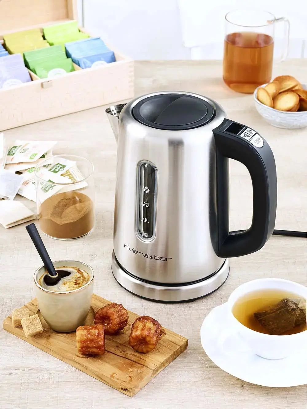 La Bouilloire Siena - Inox (1 L) de riviera&bar est posée sur une table en bois avec une tasse de café, un thé, un sucrier, des muffins, des sachets de thé, et un verre de thé avec des biscuits en arrière-plan.