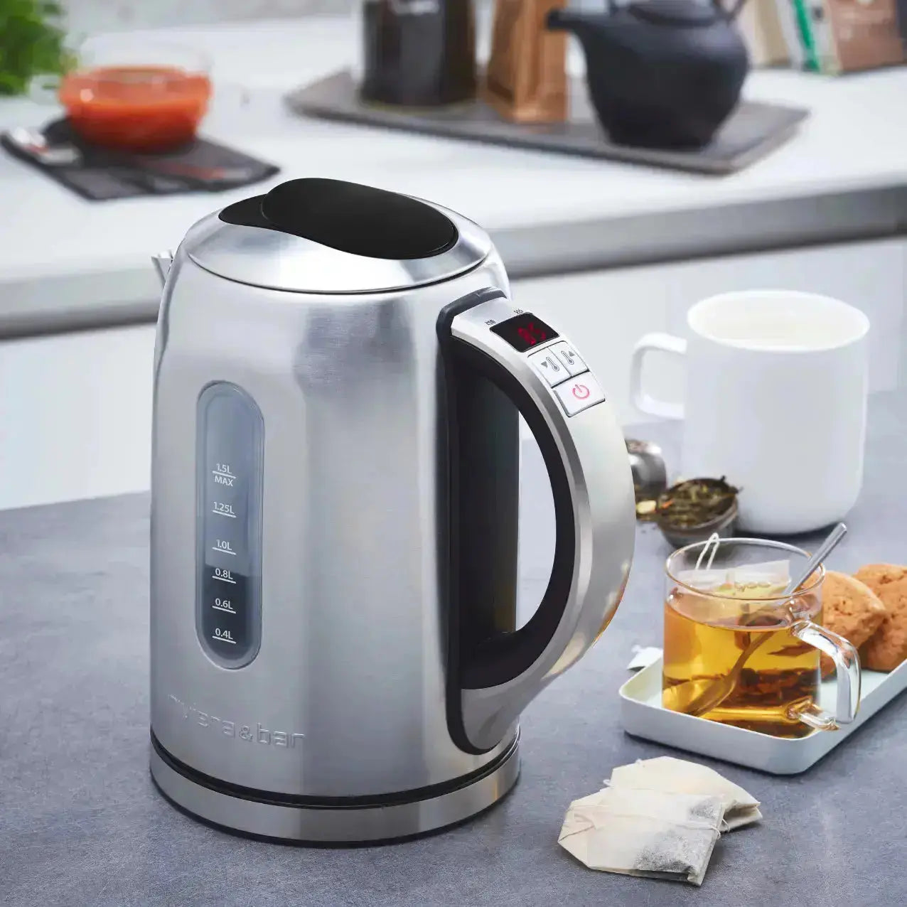 Une Bouilloire Mila - Inox (1,5 L) de riviera&bar avec affichage digital et thermostat réglable est posée sur le comptoir d'une cuisine à côté d'une tasse de thé, de sachets de thé, d'une passoire et de biscuits ; une théière et une casserole se confondent à l'arrière-plan.