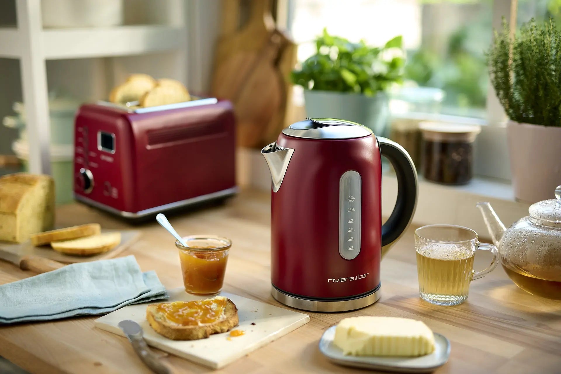 Une Bouilloire Mila - Deep Red (1.5 L) par riviera&bar est posée sur le comptoir à côté d'un grille-pain assorti, d'une théière, de marmelade et de toasts beurrés, son thermostat Mila assurant une température parfaite alors que la lumière du soleil passe à travers la fenêtre.