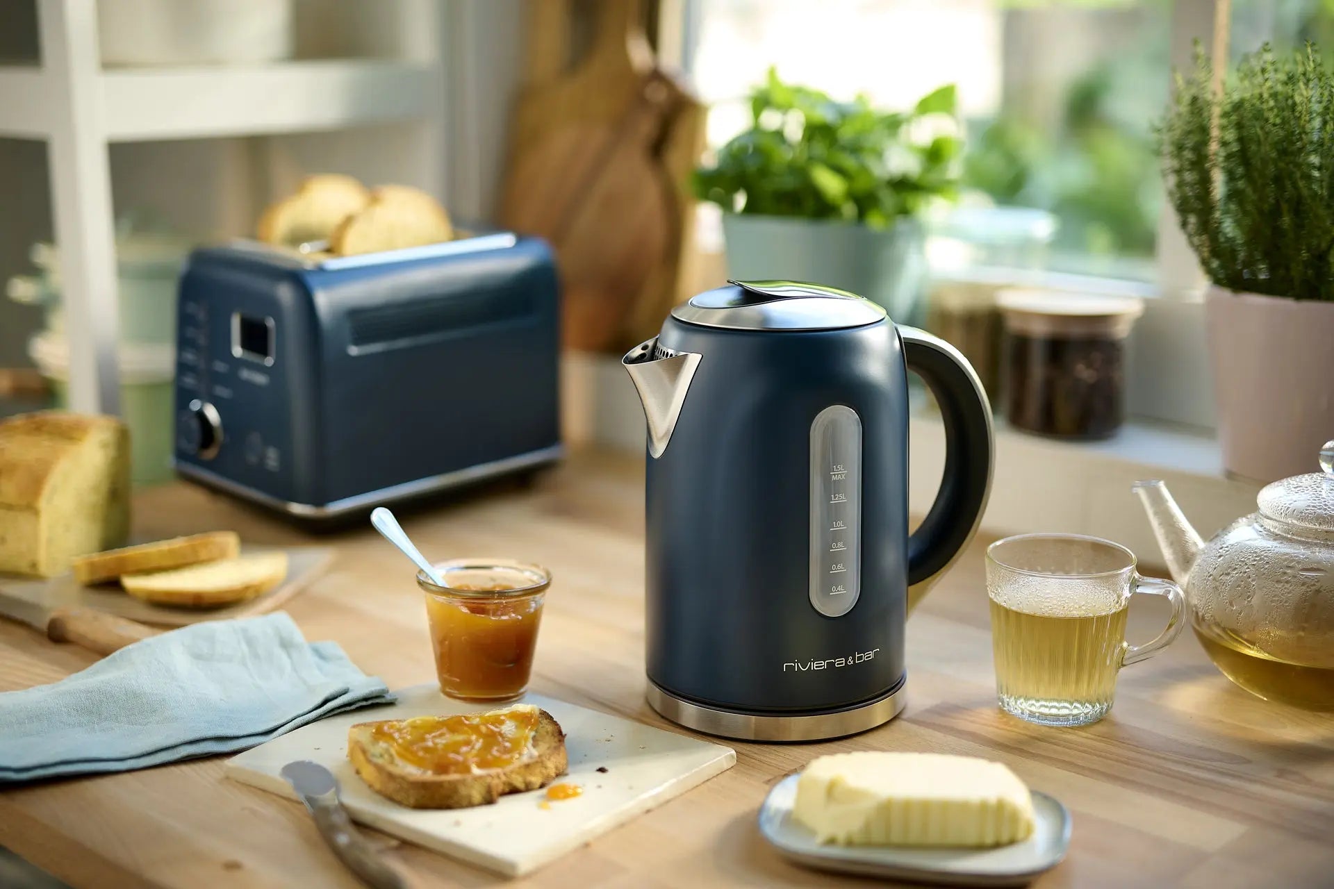 Une Bouilloire Mila - Grey Blue (1.5 L) par riviera&bar est posée sur le comptoir d'une cuisine à côté d'un grille-pain, de thé, de beurre, d'un verre à thé, de pain, d'un pot de marmelade, et de toasts avec de la marmelade alors que la lumière du soleil coule à travers la fenêtre.