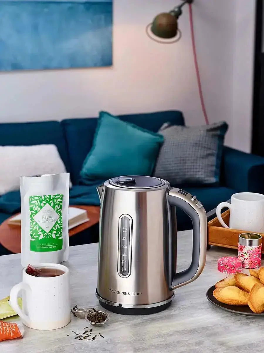 Une Bouilloire riviera&bar Elea - Inox (1,8 L) repose sur une table en marbre à côté de thé, de biscuits et de mugs. Un canapé bleu avec des coussins et une lampe en arrière-plan ajoutent une touche de confort.