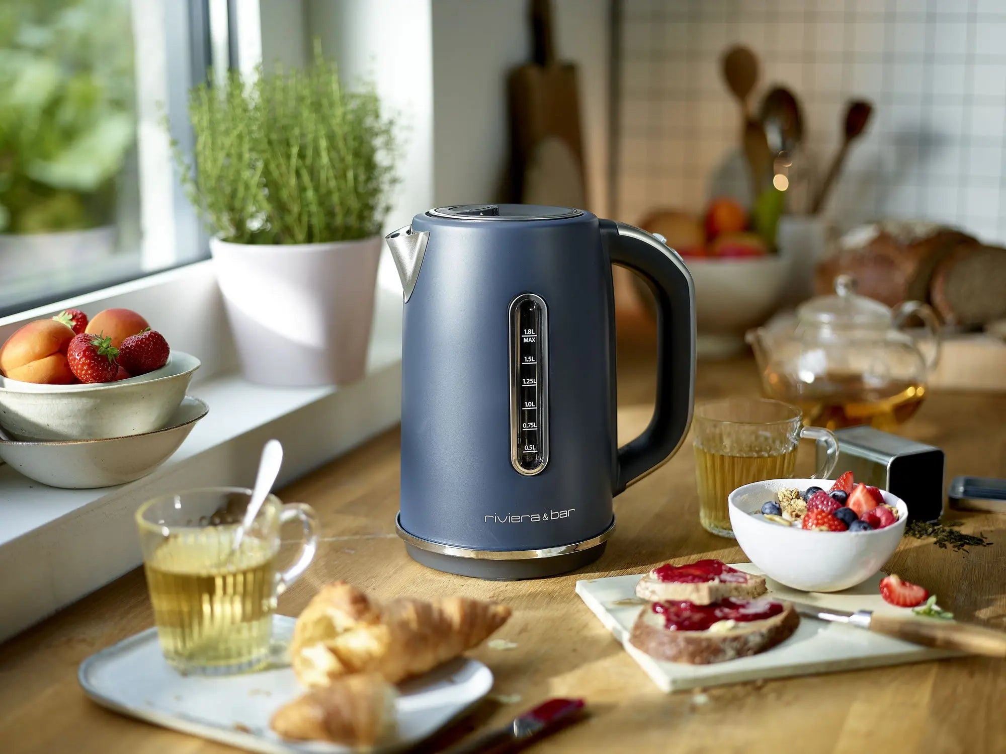 Une riviera&bar Bouilloire Aria - Grey Blue (1,8 L) repose sur une table de cuisine en bois avec des tasses de thé, des croissants, des toasts avec de la confiture et du pain frais. En arrière-plan, une herbe en pot et une coupe de fruits ajoutent de la couleur près de la fenêtre.
